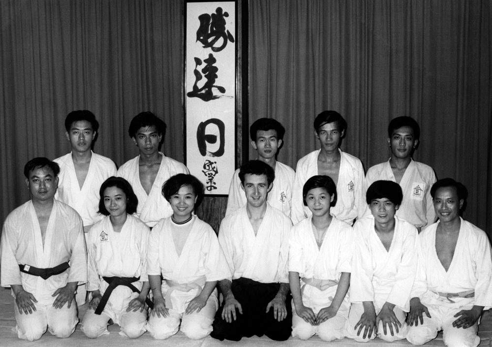 Alan Ruddock posant avec les élèves de l'Aikikai de Hong Kong devant une calligraphie donnée par O Sensei à Virginia Mayhew. Les caractères 勝速日 font partie de l'une des expressions préférées du fondateur du Kojiki : 正勝吾勝勝速日.